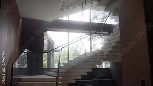 Modern floating staircase in bright atrium with backlit windows and glass balustrade, low mid-level upward viewpoint emphasizing diagonal geometry and minimalist mood