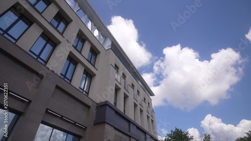 Low-angle tilt of modern mid-rise office building corner under blue sky with scattered cumulus clouds, emphasizing vertical lines and rooftop terrace with building, architecture, facade, office