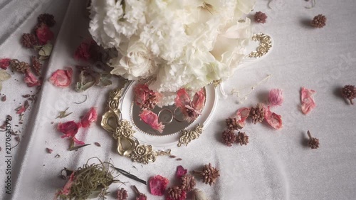 Ornate gold-accented handheld mirror reflecting two rings beside white bouquet, dried pink petals and seed pods on pale fabric,elegant floral jewelry arrangement