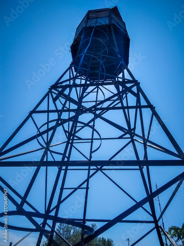 Quadro su tela Metal water tower structure rising against a clear bright blue summer sky photog