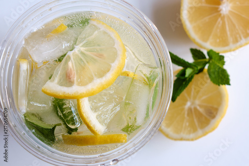 An overhead close-up shot of a clear takeaway cup with cold lemonade, ice cubes, and lemon slices next to fresh mint for beverage advertising and summer menu designs.