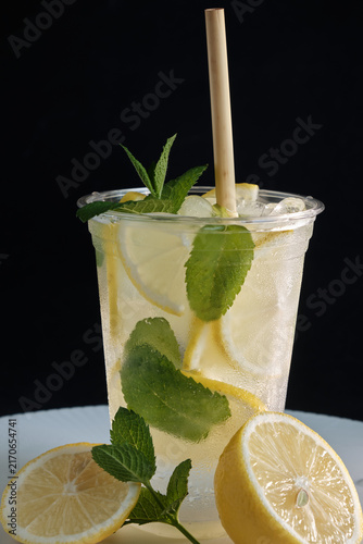 A vertical shot of a cold takeaway cup filled with iced lemonade, lemon slices, green mint leaves, and a eco straw on a dark background for beverage branding and social media ads.
