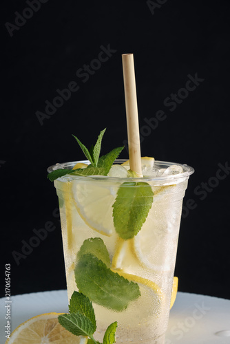 A vertical shot of a cold takeaway cup filled with iced lemonade, lemon slices, green mint leaves, and a eco straw on a dark background for beverage branding and social media ads.