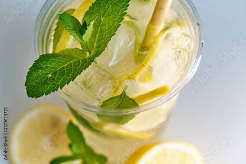 An overhead close-up shot of a clear takeaway cup with cold lemonade, ice, lemon slices, mint, and a straw for beverage advertising and summer menu promotions.