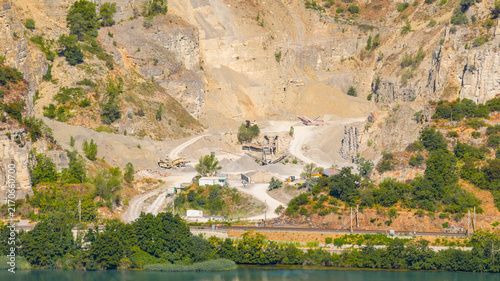 Aerial view of an open pit quarry featuring exposed rock faces, aggregate stockpiles, and heavy crushing equipment with haul roads in Le Pouzin, Auvergne-Rhone-Alpes, France.