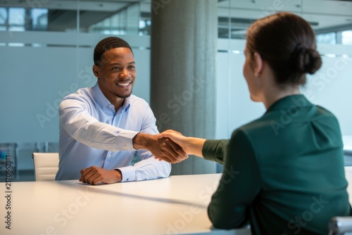 A friendly african american man in a light blue shirt shaking hands with a woman in a green blazer during a job interview in a bright, contemporary office environment