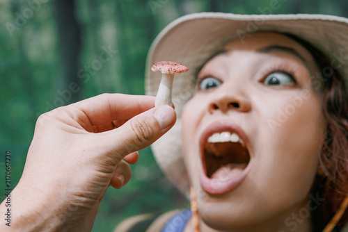 Foto Wide-eyed woman in sun hat funny eating tiny wild mushroom close to her open mouth