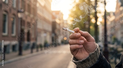 Person holding a lit cigarette while standing on a European city street at sunset with soft glowing golden sunlight in the background