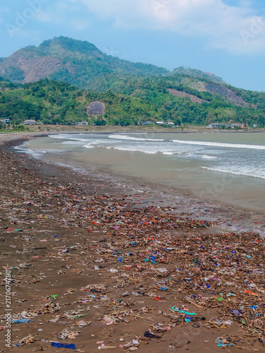 Aerial view of a beach heavily polluted with plastic waste and debris, with rolling waves and lush green mountains in the background, Indonesia.