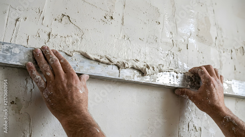 Mason leveling fresh plaster on a wall surface using a long aluminum straight edge tool.