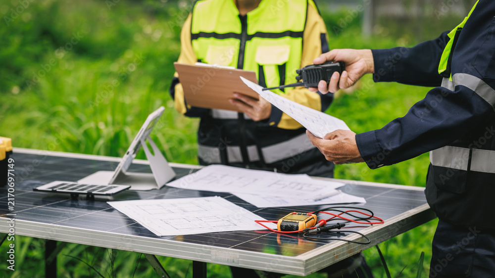 Fototapeta premium Engineers in safety vests review plans and use a radio beside solar panels and electrical testing tools at an outdoor renewable energy site.