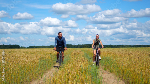Konstfotografi Front view of a smiling man and woman in athletic wear on a bicycle trip, riding along a countryside route in a vast wheat field during a peaceful summer weekend