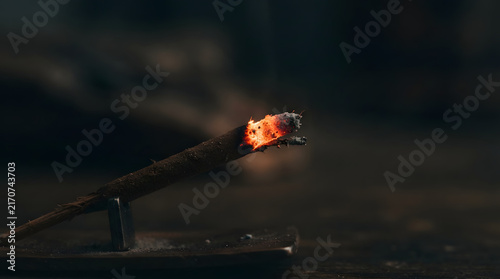 Incense stick burning with a glowing tip and smoke on a small metal holder against a dark background