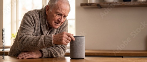 A close-up of a thoughtful elderly man in a warm cardigan leaning on a wooden kitchen counter and touching a modern voice-controlled smart speaker device.