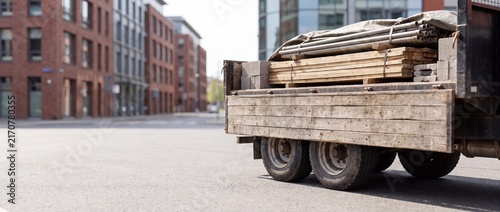 A close-up view of the back of a wooden flatbed truck loaded with timber, pipes, and bricks covered by a tarp, parked on an empty city street.