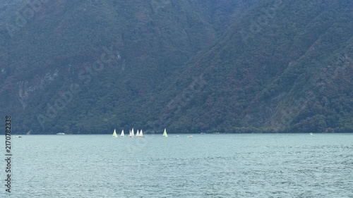 Several small sailboats navigate across a tranquil, pale blue lake, with a majestic, tree-covered mountain range forming a dramatic backdrop under an overcast sky. Switzerland, Lugano, Lake Lugano