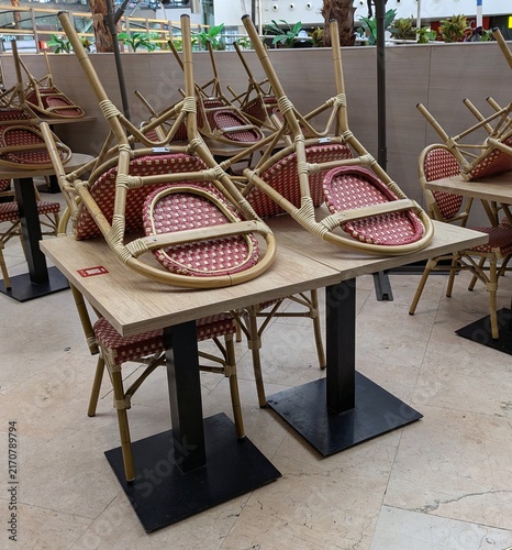 Mesa de madera con sillas estilo bistró con asiento tejido en rojo y blanco colocadas boca abajo, en un restaurante cerrado 