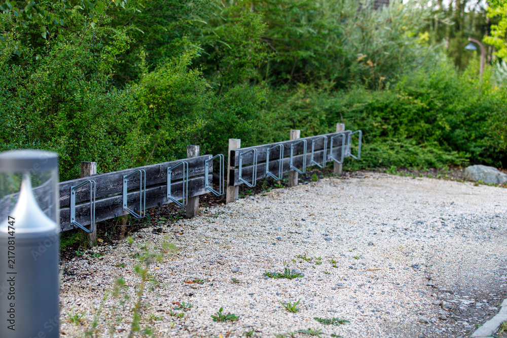 Fototapeta premium An empty wooden bicycle rack with metal holders sits beside a gravel path in a lush green park.