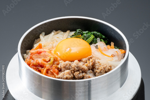 Korean bibimbap served in a traditional stone bowl, topped with a bright yellow egg, ground pork, fresh vegetables, and bean sprouts arranged in an appetizing presentation