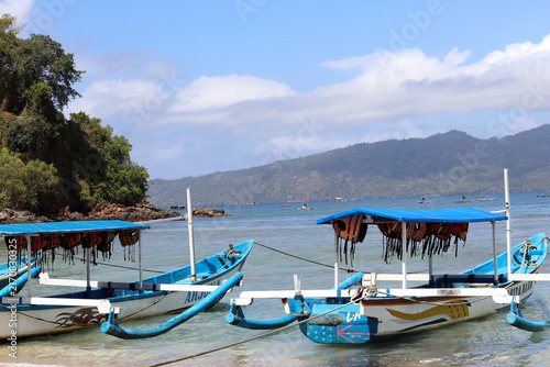 Traditional wooden boats rest on a sandy beach facing clear waters, surrounded by lush tropical mountains under a cloudy sky.