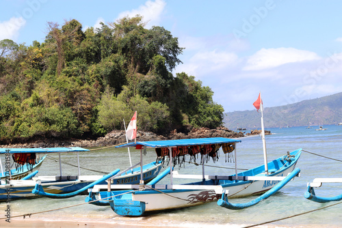 Traditional wooden boats rest on a sandy beach facing clear waters, surrounded by lush tropical mountains under a cloudy sky.