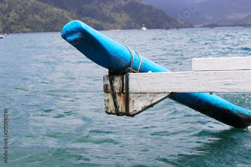 A close-up view of a bright blue wooden outrigger float tied with rope to a  support arm on a boat, set against calm ocean waters and distant hills.
