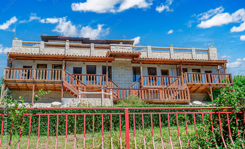 Fototapeta premium A two-story stone house features wooden balconies under a bright blue sky filled with white clouds.
