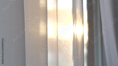 Handheld camera moving past sheer white curtains glowing in warm evening sunlight as the fabric gently sways in a summer breeze.
