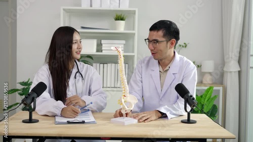Asian female doctor pointing at a spine model explaining orthopedics to a male doctor during a medical podcast interview. Recording a healthcare audio broadcast in a modern clinic studio.