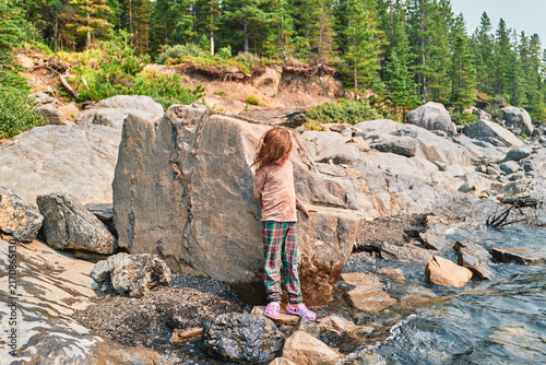 Boy standing beside a large boulder on a rocky lakeshore from behind
