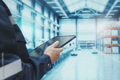 A close-up shot shows a factory worker using their finger to control a digital tablet. The backdrop is a large, modern manufacturing plant with automated production lines, shelving, control systems, a