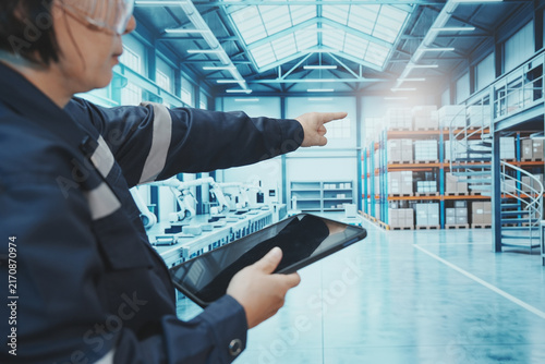 A close-up shot shows a factory worker using their finger to control a digital tablet. The backdrop is a large, modern manufacturing plant with automated production lines, shelving, control systems, a
