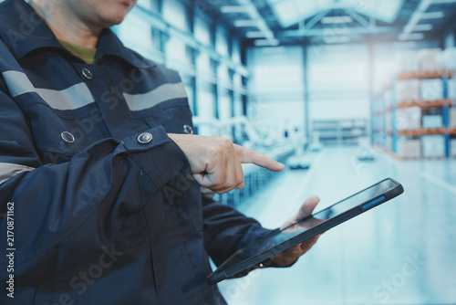 A close-up shot shows a factory worker using their finger to control a digital tablet. The backdrop is a large, modern manufacturing plant with automated production lines, shelving, control systems, a