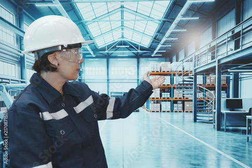 A close-up shot shows a factory worker using their finger to control a digital tablet. The backdrop is a large, modern manufacturing plant with automated production lines, shelving, control systems, a