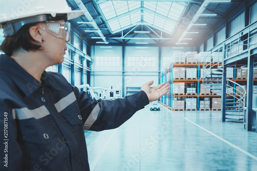 A close-up shot shows a factory worker using their finger to control a digital tablet. The backdrop is a large, modern manufacturing plant with automated production lines, shelving, control systems, a