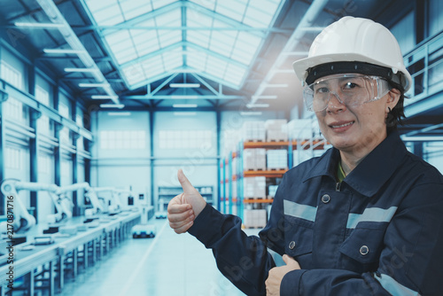 A close-up shot shows a factory worker using their finger to control a digital tablet. The backdrop is a large, modern manufacturing plant with automated production lines, shelving, control systems, a