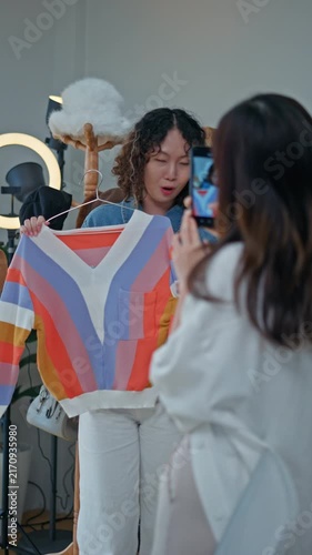 Young Asian woman fashion entrepreneur live streaming selling clothes online with smartphone ring light and laptop in home studio, representing small business owner and professional women.