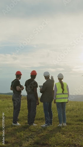 Diverse construction workers in hard hats and vests stand on grassy field discussing plans with tablet Suitable for technology, engineering and renewable energy content.