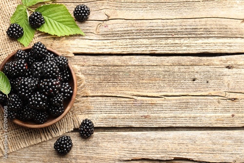 Ripe blackberries in bowl with leaves on wooden table, flat lay. Space for text