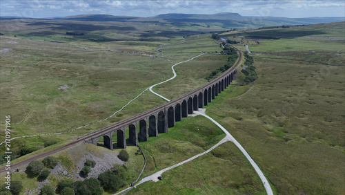 Aerial view of the historic Ribblehead Viaduct spanning the vast green landscape of the Yorkshire Dales with railway tracks and winding paths in Carnforth, England, United Kingdom.