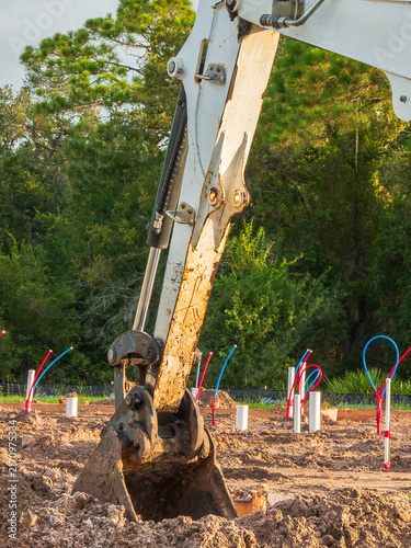 Bucket on a compact excavator digs into sandy ground, maybe to dig a trench, near a group of utility conduits for house construction on a lot in a suburban residential development in southwest Florida