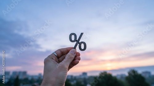 Hand holding a wooden percent symbol against a pink and orange sunset sky