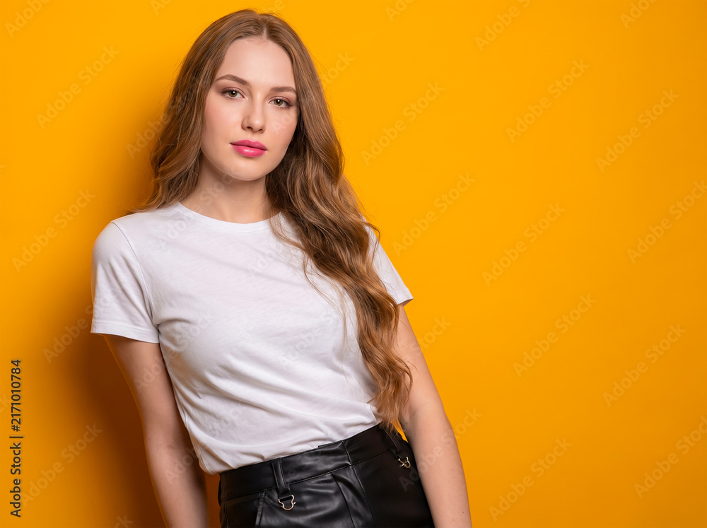 Fototapeta premium Beautiful young woman portrait in blank white t-shirt and black leather skirt posing against a bright yellow background with copy space for casual fashion mockup.