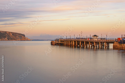 A clear November evening long exposure looking across smooth waters along Swanage Pier with a pink sky