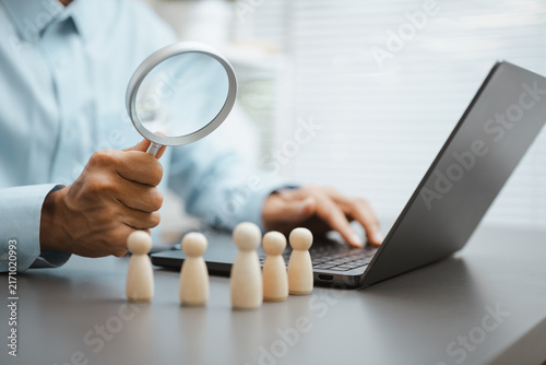 Фотография A recruiter holds a magnifying glass over wooden people figures beside a laptop while comparing each candidate for a hiring decision and planning the next recruitment interview for the open position