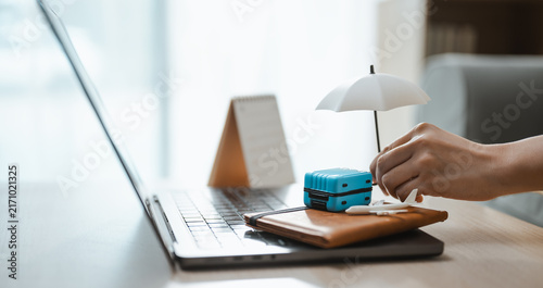 Foto A hand arranges a suitcase and airplane beneath an umbrella beside a laptop to compare travel insurance coverage for medical care and lost luggage before an overseas journey or unexpected delay