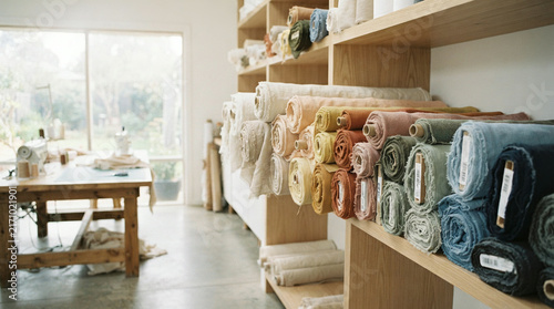 Colorful rolls of fabric neatly arranged on wooden shelves in a sunlit textile workshop or studio