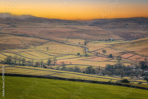 A scenic view of rolling countryside, dry stone walls and winding lanes beneath a golden sunset near Malham, a small village in Yorkshire Dales, UK