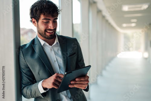 Принти на полотні Young businessman smiling, leaning against a wall in a modern office, working wi