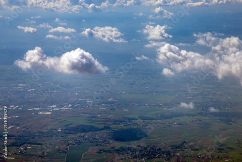An aerial view from an airplane window looking down at fields, towns, and scattered cloud formations under a bright blue sky. The landscape below displays a patchwork of agricultural fields and roads.